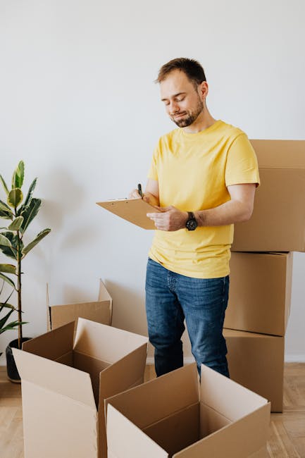A person wearing a yellow t-shirt and blue jeans is inside a room, engaged in a home relocation or furniture transport process, holding a clipboard with a pen as they prepare to record details during 